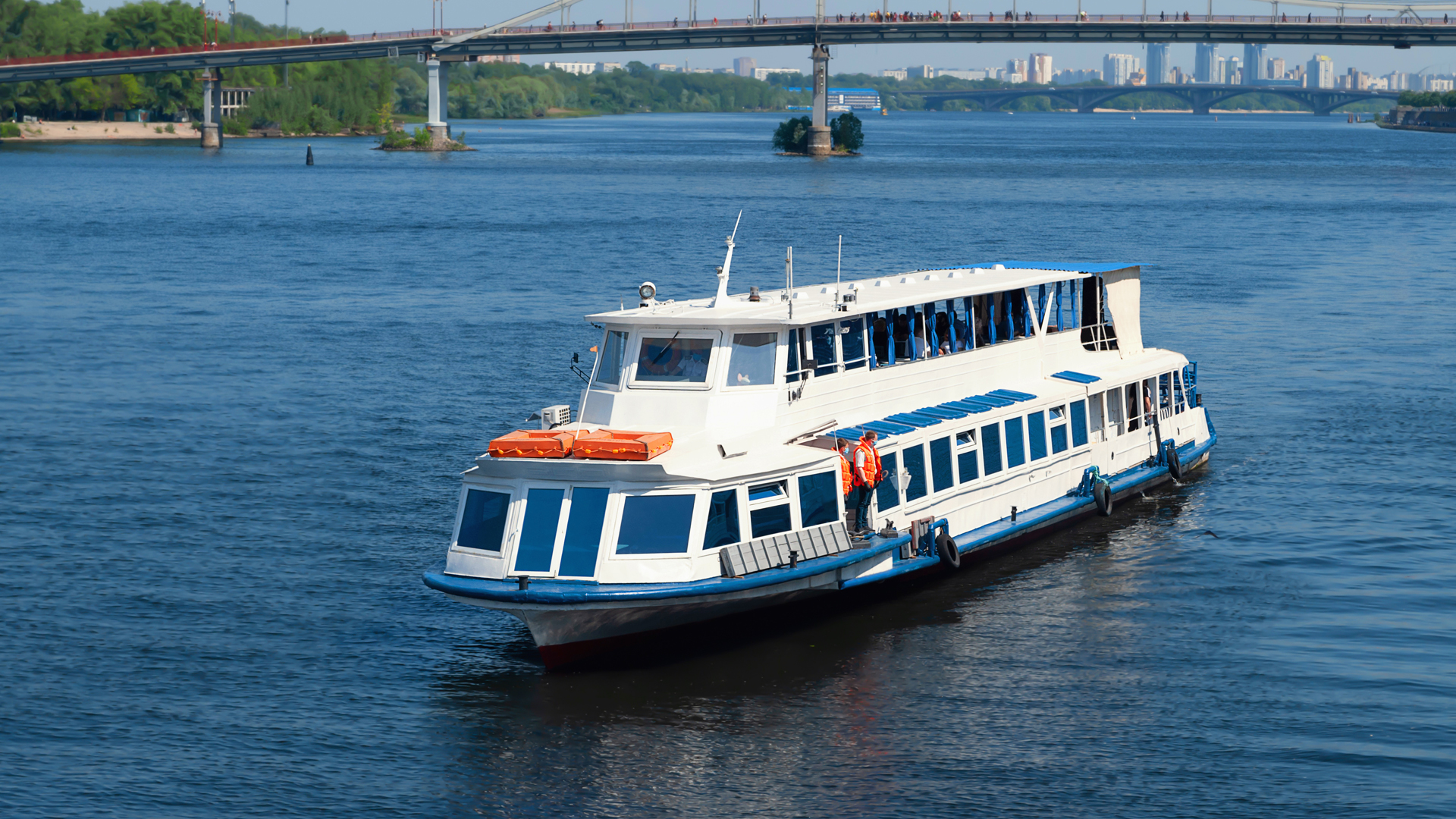 Blue-and-white excursion boat on a wide river below a long bridge